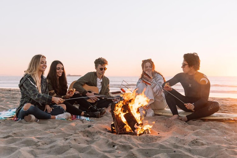Group of people sitting on the beach with a bonfire during sunset.