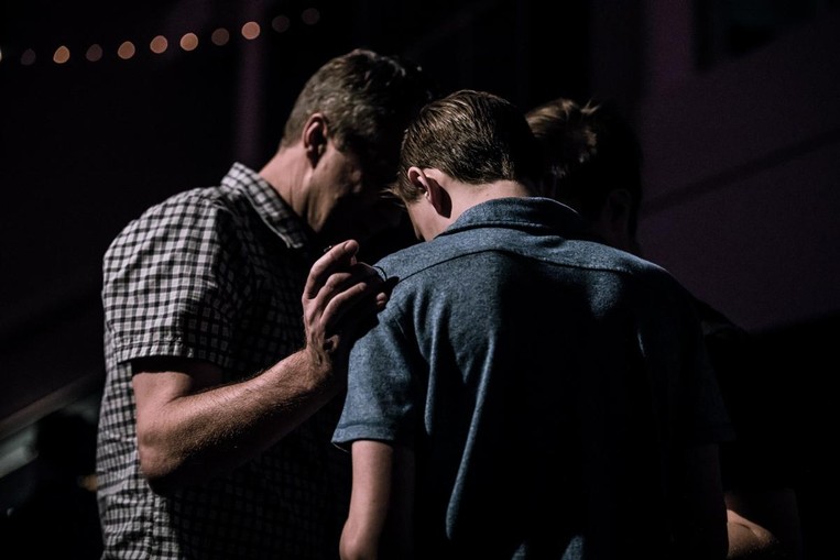 A man and his son pray together in a dark room. The father is laying a hand on his son's shoulder.