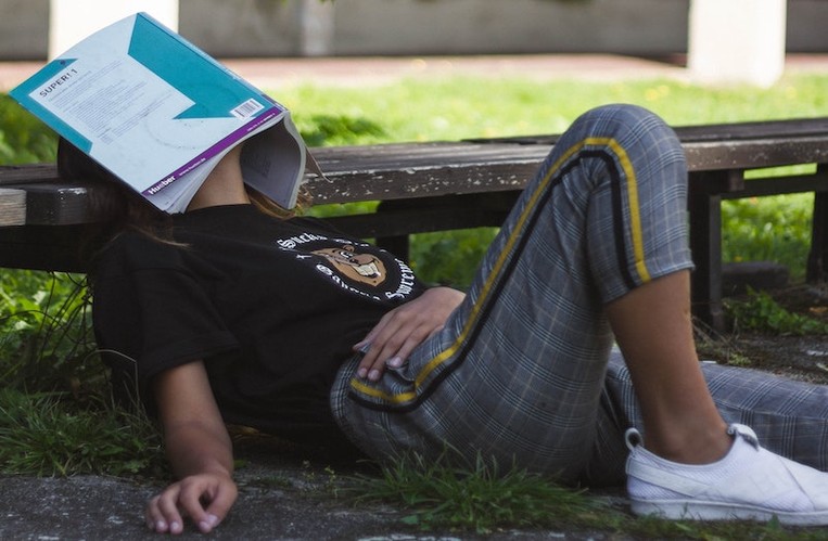 A girl is sitting on the grass with a textbook over her face.
