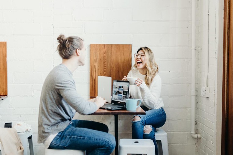 A guy and a girl sit at a table together with their computers laughing.