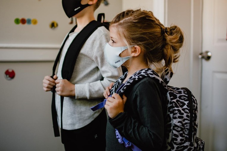 Two kids, a boy and a girl, stand in a classroom with their backpacks on and with masks on.