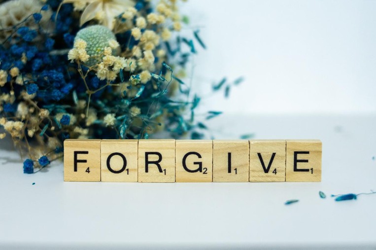 Scrabble game tiles spell out the word "Forgive" with blue bouquet of flowers behind it. 