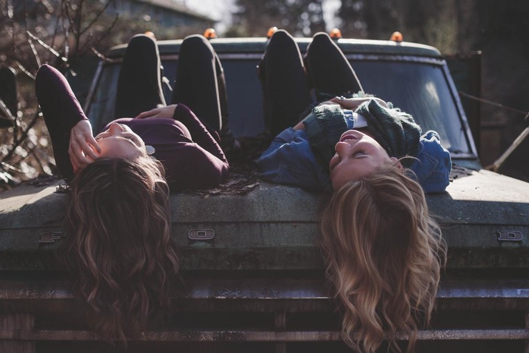 Two girls lay on top of a car with space between them.